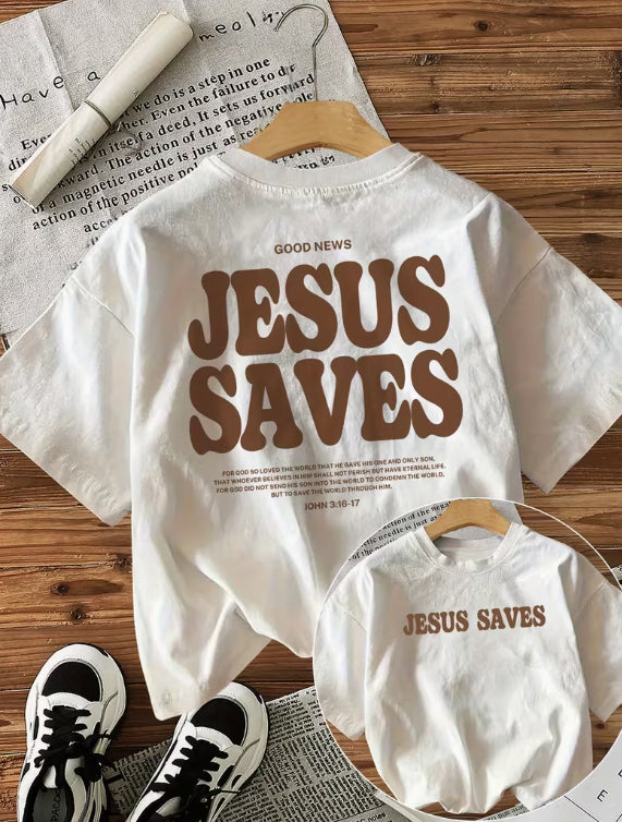 White t-shirt with 'Jesus Saves' text on a wooden surface with a baby onesie and sneakers.