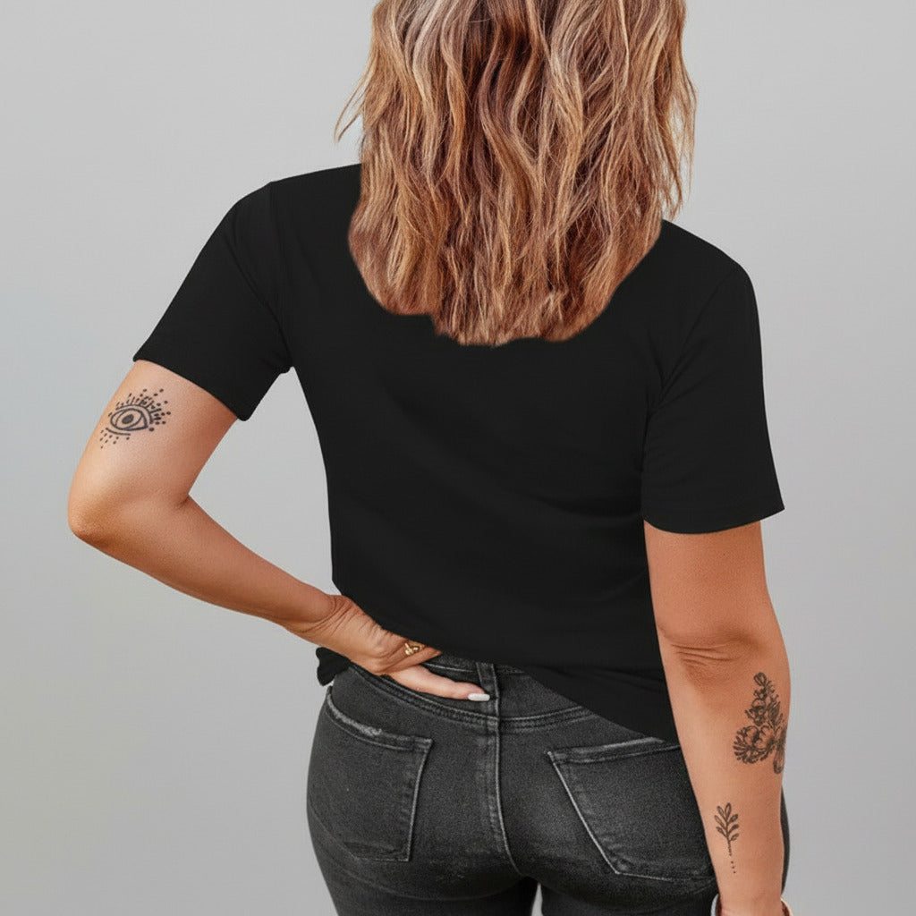 Person wearing a black t-shirt and dark jeans with tattoos on arms against a stone wall.